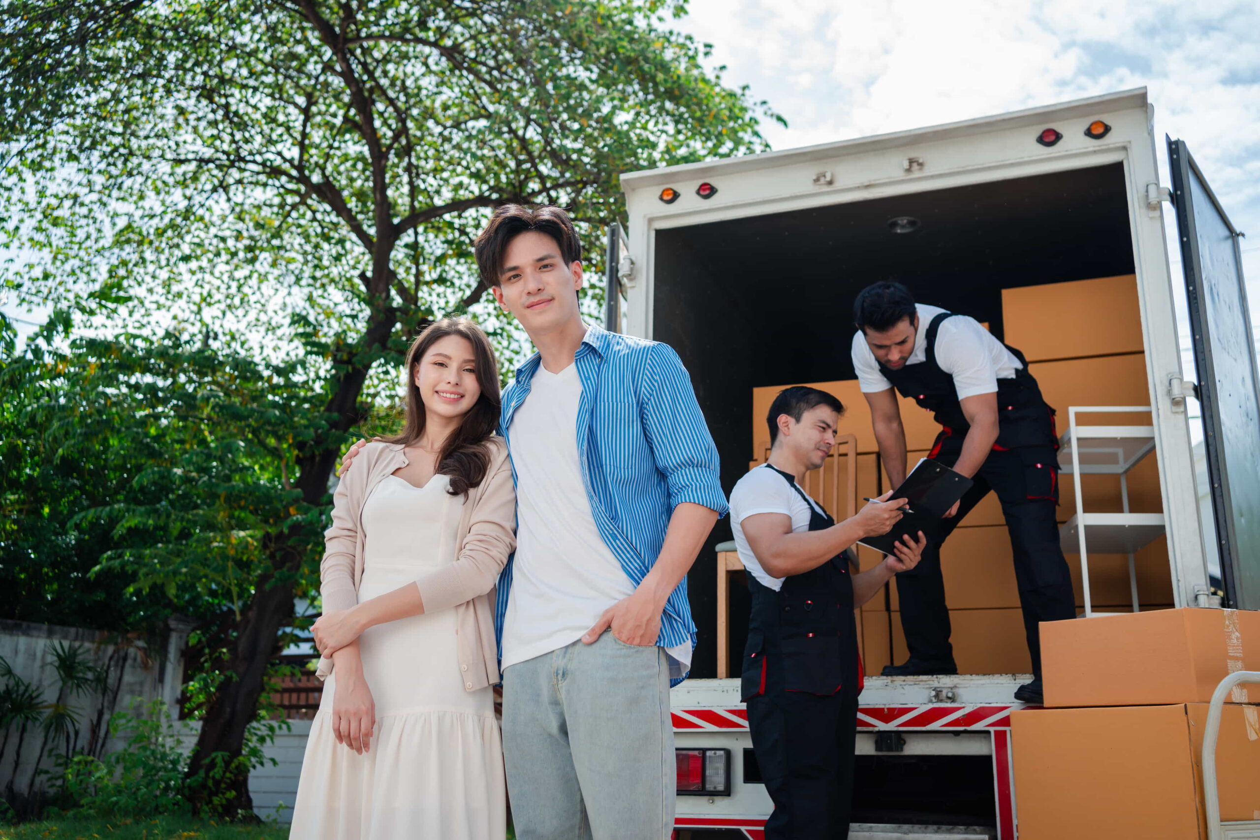 A Couple standing together while smiling with movers and a moving truck behind them