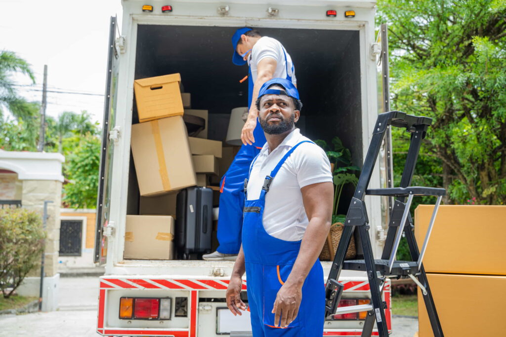  Two movers wearing blue overalls unloading packing boxes from a moving truck.